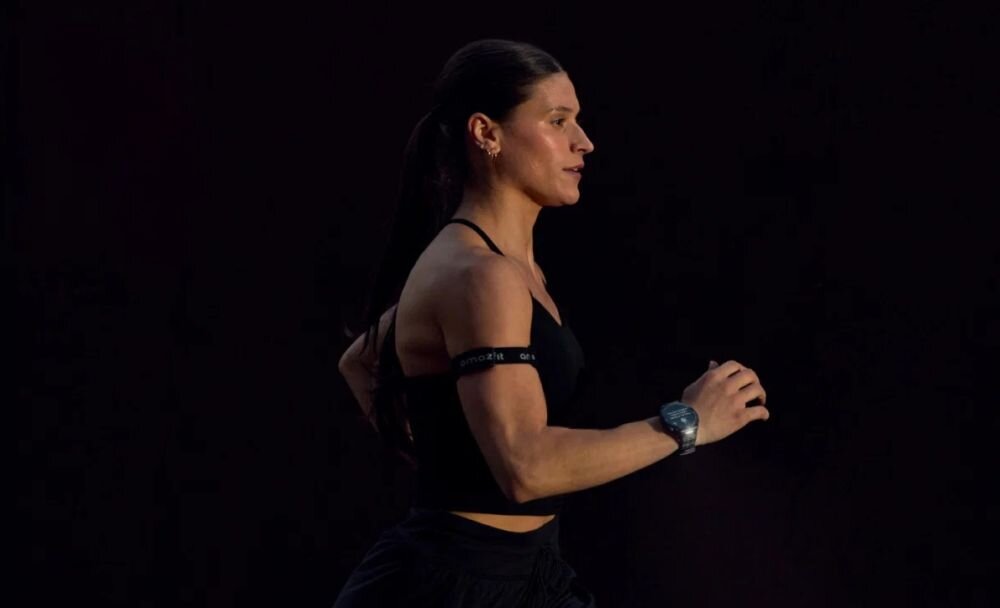Osoba biegnąca w ciemnym stroju sportowym, na nadgarstku widoczny smartwatch. Tło jest ciemne, co podkreśla sylwetkę biegacza.