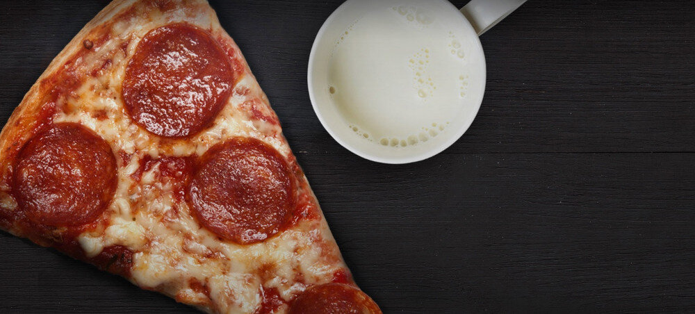 KUCHNIA MIKROFALOWA LG MS23NECBW Zbliżenie na duży trójkątny kawałek pizzy pepperoni na ciemnym drewnianym tle, obok którego stoi biała filiżanka z mlekiem, tworząc kontrastowe, apetyczne ujęcie potrawy.