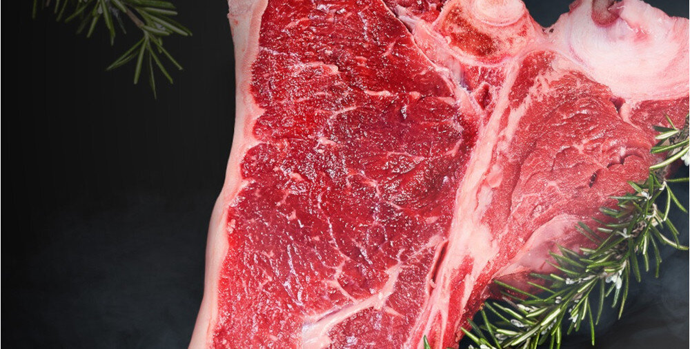 KUCHNIA MIKROFALOWA LG MS23NECBW Zbliżenie na surowy, marmurkowany kawałek czerwonego mięsa z kością, udekorowany gałązkami rozmarynu na ciemnym tle, podkreślające świeżość i strukturę steka.