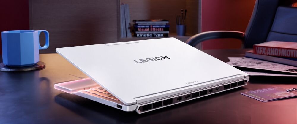 Na biurku znajduje się biały laptop z napisem \'LEGION\' na pokrywie. W tle widoczny jest kubek oraz kilka książek i dokumentów.
