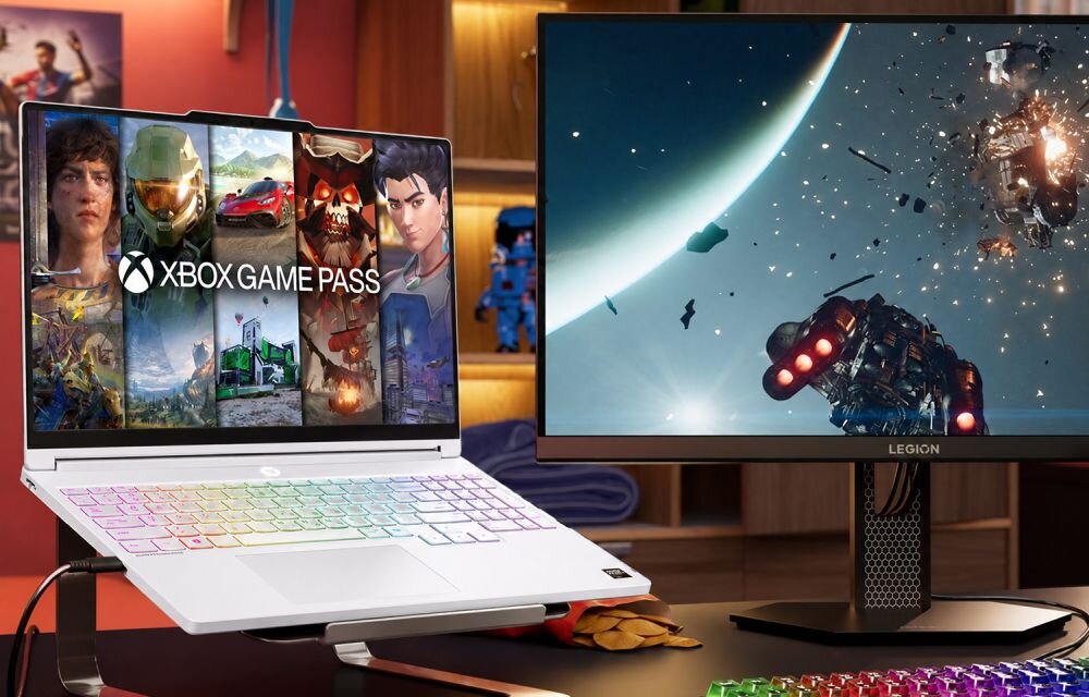 Na obrazie widać laptop z kolorową klawiaturą i ekranem wyświetlającym grafikę z napisem \'XBOX GAME PASS\'. Obok znajduje się monitor z logo \'LEGION\', na którym widoczna jest scena kosmiczna z eksplozją.