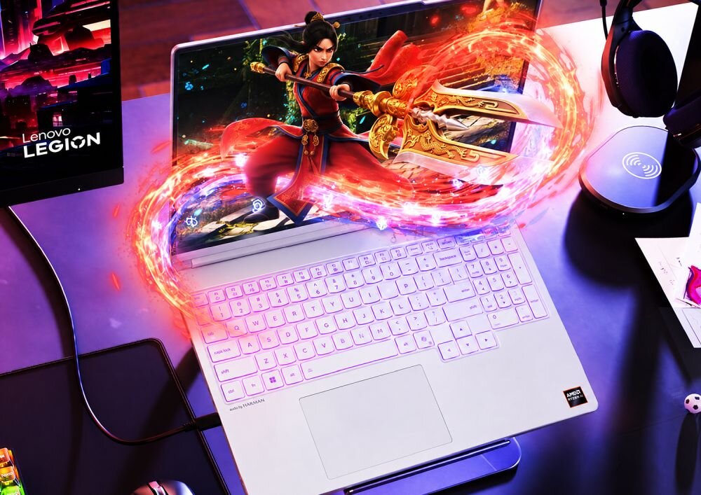 Na obrazie znajduje się biały laptop z podświetlaną klawiaturą, na ekranie którego widoczna jest postać z gry w dynamicznej pozie. Obok laptopa znajduje się monitor z logo \'Lenovo LEGION\'.