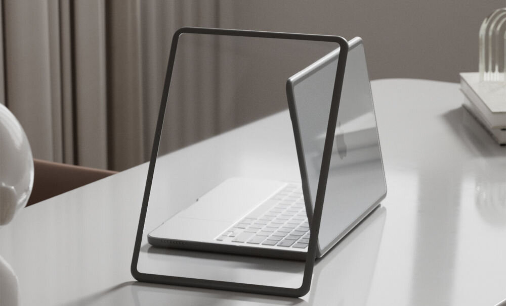 Na białym stole znajduje się otwarty laptop, a obok niego przezroczysto-czarny element przypominający ramkę. W tle widoczne są zasłony i kilka książek.
