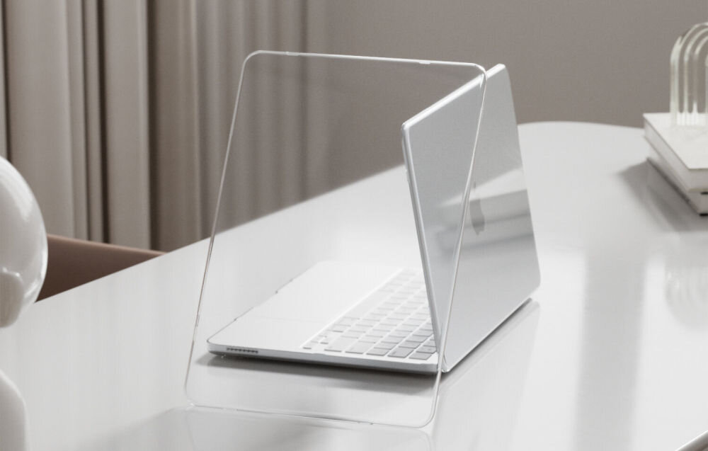 Na białym stole znajduje się otwarty laptop z przezroczystym etui. W tle widoczne są zasłony i stos książek.