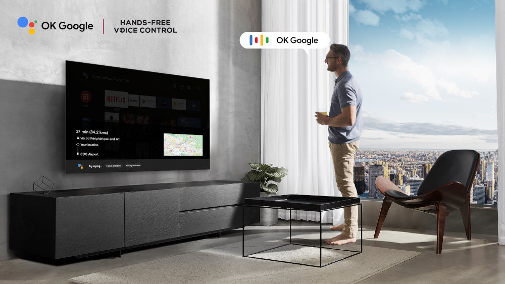 W salonie na ścianie wisi telewizor z włączonym interfejsem Google TV, obok stoi mężczyzna. Na ekranie widoczne są aplikacje oraz mapa, a na ścianie logo \'OK Google\' i \'HANDS-FREE VOICE CONTROL\'.