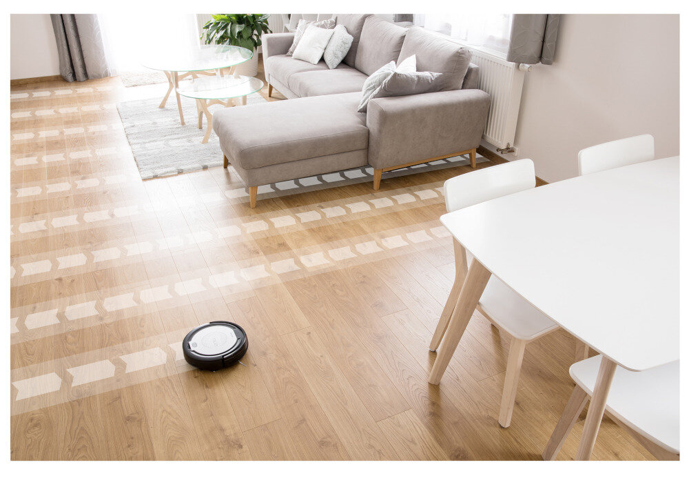 W jasnym salonie z drewnianą podłogą znajduje się robot sprzątający, który porusza się po wyznaczonej trasie. W tle widoczna jest szara sofa, stolik kawowy i biały stół z krzesłami.