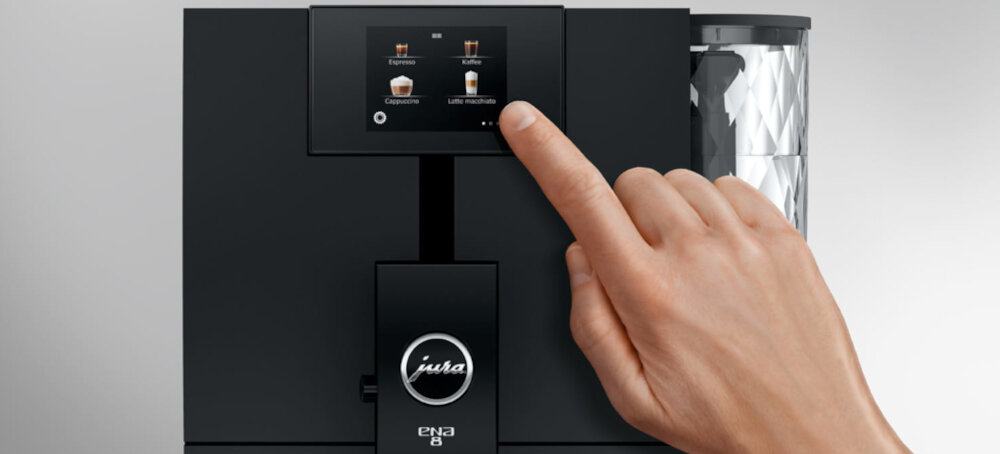 Osoba dotyka ekranu dotykowego na czarnym ekspresie do kawy z widocznym logo \'jura\' oraz napisem \'ena 8\'. Na ekranie wyświetlane są opcje napojów: Espresso, Coffee, Cappuccino, Latte macchiato.