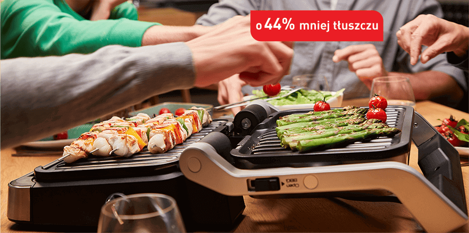 Na stole znajduje się grill elektryczny z potrawami, takimi jak szaszłyki i szparagi. Widoczny jest napis \'o 44% mniej tłuszczu\'.