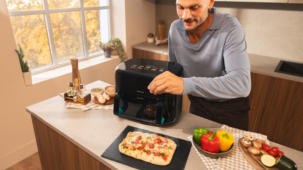 Na blacie kuchennym stoi frytkownica beztłuszczowa z widocznym panelem sterowania. Obok leży pizza oraz kolorowe warzywa na desce do krojenia.
