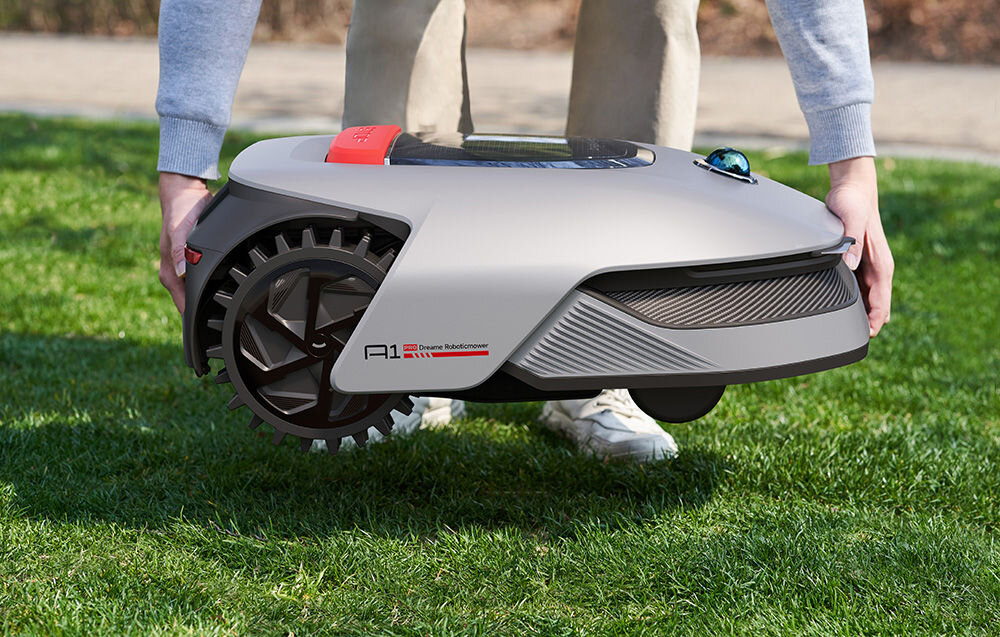 Osoba trzyma szary robot koszący z widocznym napisem \'A1 Dreame RoboticMower\' na trawie. Robot ma duże koła i nowoczesny design.