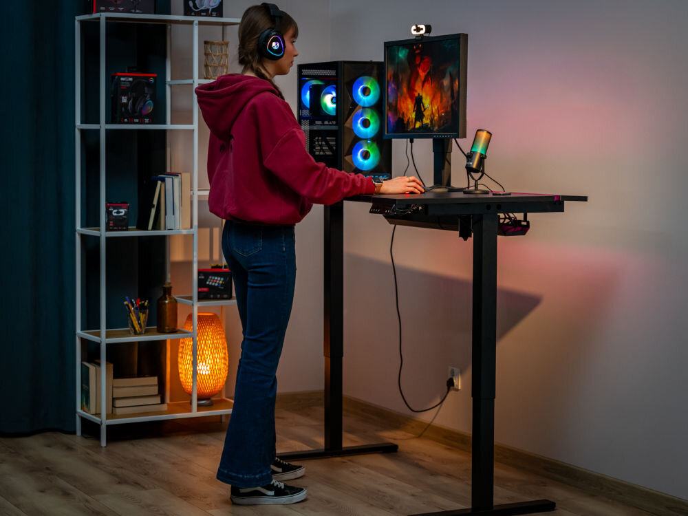 Osoba stoi przy elektrycznym biurku regulowanym z komputerem i monitorem, na którym wyświetlana jest kolorowa grafika. W tle znajduje się regał z książkami i lampka podłogowa.