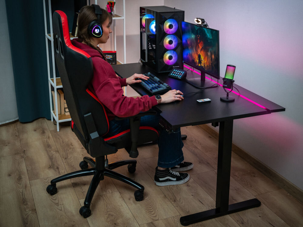 Osoba siedzi przy czarnym biurku elektrycznym z regulacją wysokości, na którym znajduje się monitor, klawiatura, myszka i komputer z podświetleniem RGB. Na biurku widoczny jest również mikrofon i kontroler do streamingu.