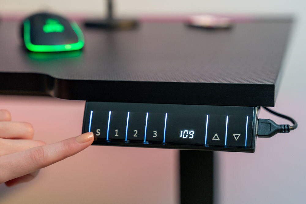 Panel sterowania biurka elektrycznego z przyciskami oznaczonymi jako S, 1, 2, 3 oraz wyświetlaczem pokazującym \'109\'. Obok panelu znajduje się mysz komputerowa z podświetleniem.