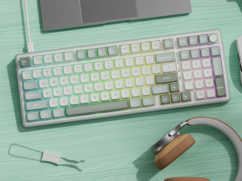 Na obrazie znajduje się klawiatura z kolorowymi klawiszami, obok której leżą słuchawki i narzędzie do wyjmowania klawiszy. Klawiatura ma układ QWERTY i numpad, a tło jest w kolorze miętowym.
