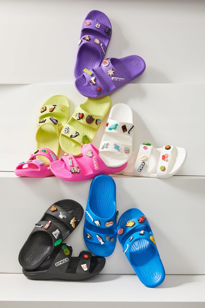 Na obrazie widoczne są różnokolorowe klapki z ozdobami, w tym fioletowe, zielone, różowe, białe, czarne i niebieskie. Na niektórych klapkach widnieje logo \'crocs\'.