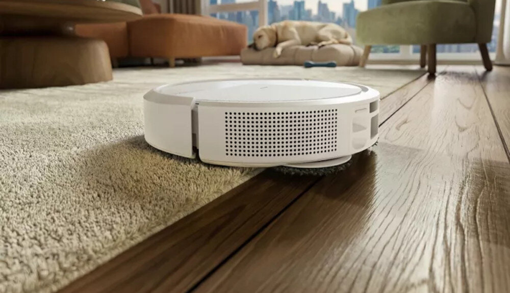 Biały robot sprzątający porusza się po podłodze, przechodząc z dywanu na drewnianą powierzchnię. W tle widać meble i psa leżącego na posłaniu.