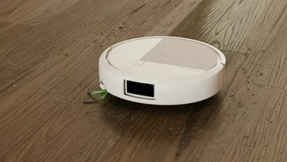 Biały robot sprzątający porusza się po drewnianej podłodze, zbierając drobne zanieczyszczenia. Na urządzeniu widoczne są przyciski i czujniki.
