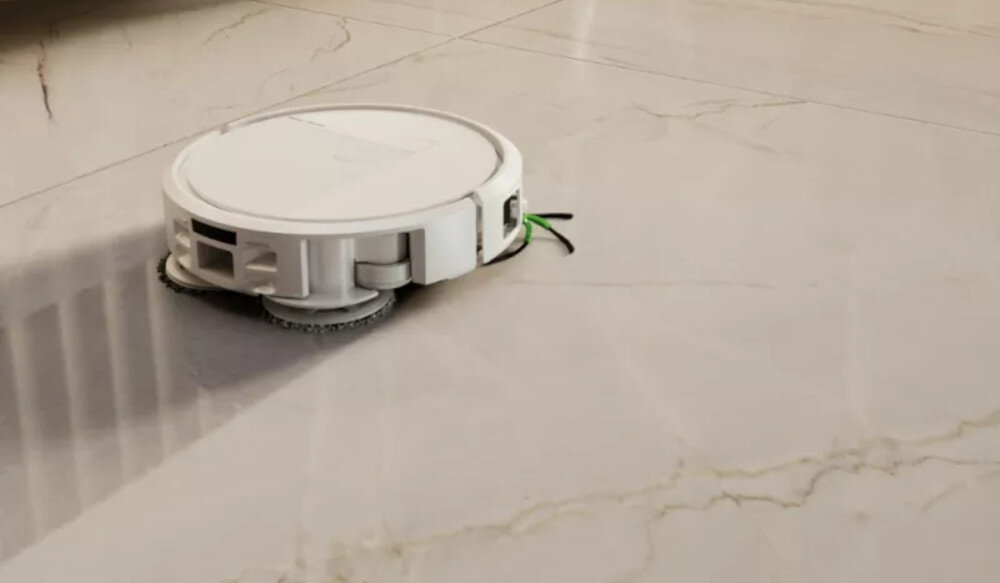 Na obrazie widoczny jest biały robot sprzątający na jasnej, marmurowej podłodze. Robot ma okrągły kształt i jest wyposażony w szczotki boczne.