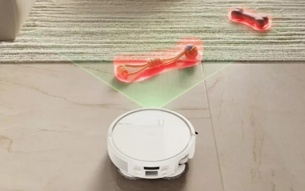 Na podłodze znajduje się biały robot sprzątający, który skanuje otoczenie zielonym światłem. Przed nim leży czerwony przedmiot na dywanie.