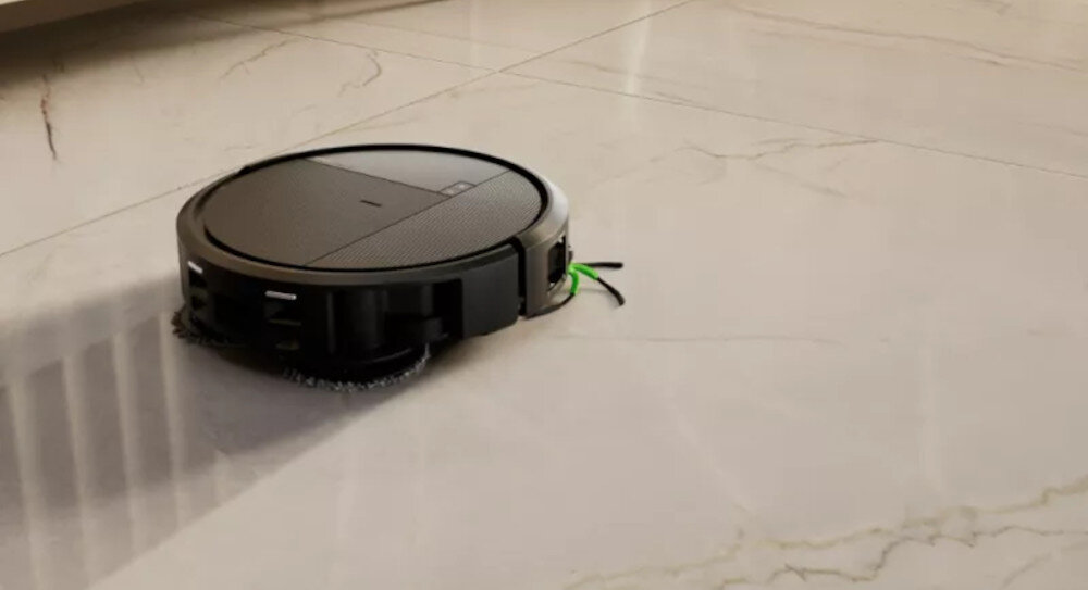 Na jasnej podłodze znajduje się czarny robot sprzątający o okrągłym kształcie z widocznymi szczotkami bocznymi.