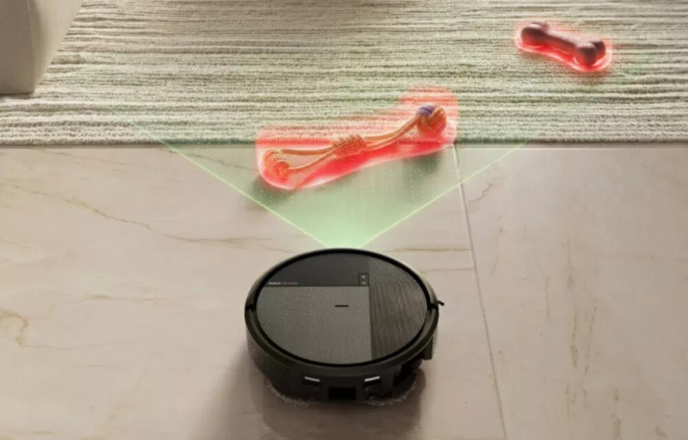 Czarny robot sprzątający porusza się po podłodze, z zielonym światłem skierowanym na czerwony przedmiot na dywanie. Na robocie widoczne jest logo \'iRobot\'.