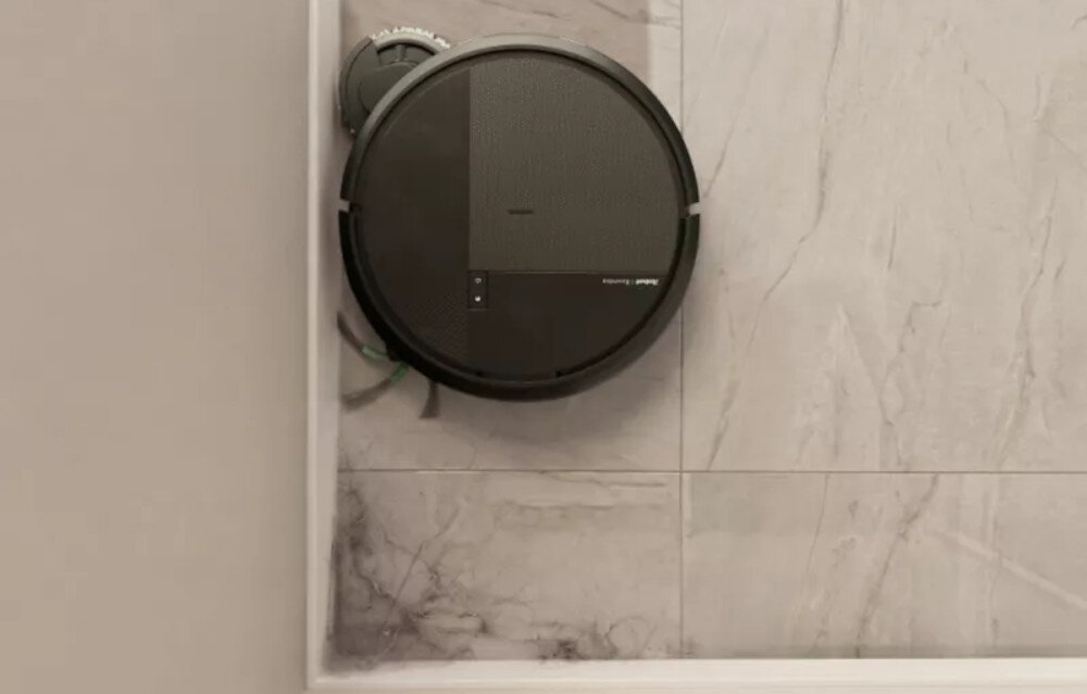 Na obrazie widoczny jest czarny robot sprzątający przymocowany do ściany w pomieszczeniu z jasnymi płytkami. Na urządzeniu znajduje się napis \'Roomba\'.