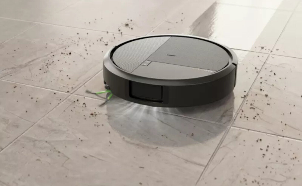 Czarny robot sprzątający na jasnej podłodze, zbierający rozsypane drobinki. Urządzenie ma okrągły kształt i widoczne są na nim elementy konstrukcyjne.