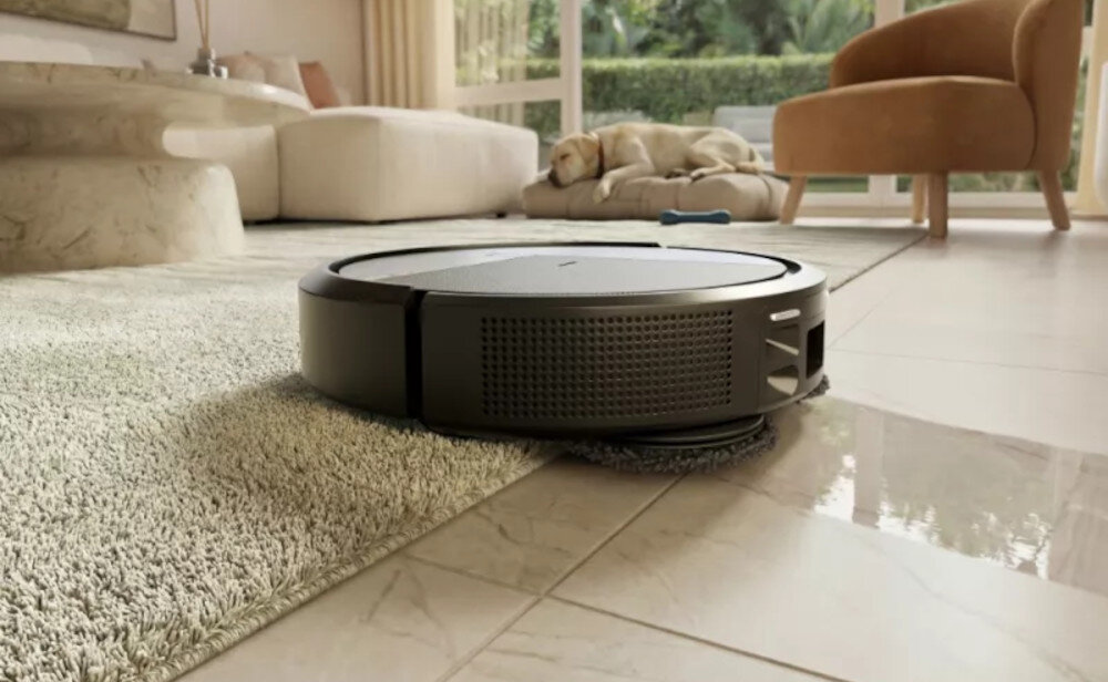 Robot sprzątający w kolorze czarnym porusza się po podłodze, częściowo na dywanie i częściowo na płytkach. W tle widać jasną sofę i psa leżącego na niej.