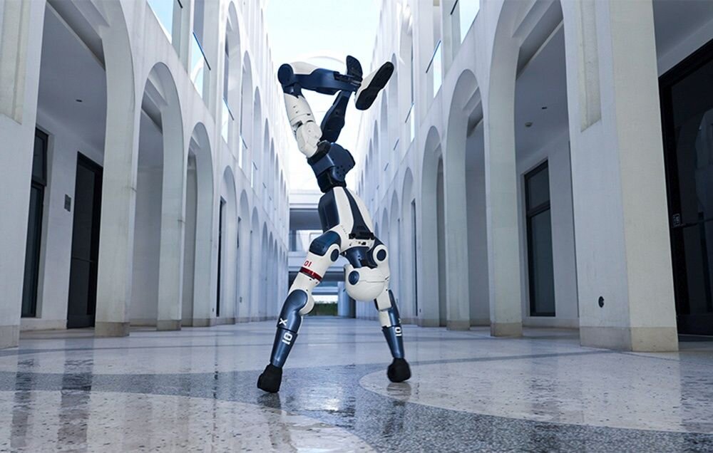 Robot humanoidalny wykonuje stanie na rękach w przestronnym korytarzu z łukowatymi przejściami. Robot ma biało-niebieską kolorystykę z widocznymi elementami mechanicznymi.