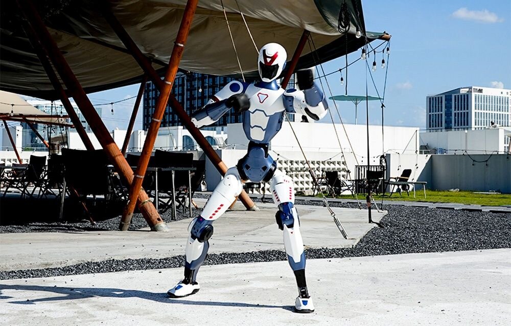 Na obrazie znajduje się humanoidalny robot w białym i niebieskim kolorze, stojący na zewnątrz pod dużym zadaszeniem. W tle widoczne są budynki i meble ogrodowe.