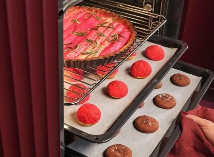 W piekarniku znajdują się dwa poziomy z jedzeniem: na górnym poziomie jest tarta z różowymi plastrami i rozmarynem, a na dolnym poziomie są czerwone i brązowe ciasteczka na papierze do pieczenia.