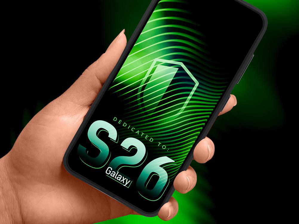 Osoba trzyma telefon z wyświetlaczem pokazującym zielone tło z falistym wzorem i symbolem tarczy. Na ekranie widoczny jest tekst: \'DEDICATED TO: S26 Galaxy\'.