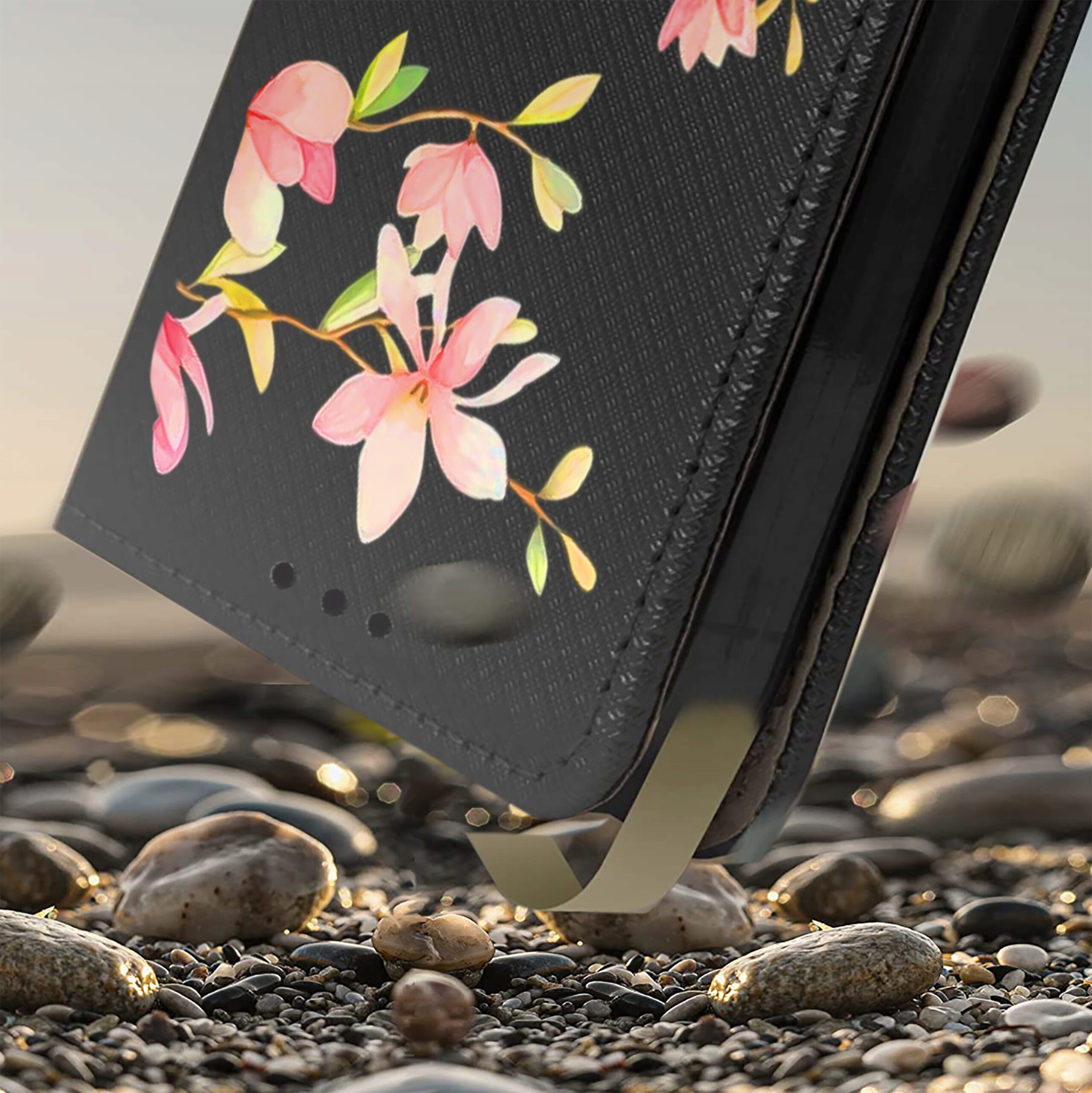 Etui na telefon z czarnym tłem ozdobione różowymi kwiatami i zielonymi liśćmi, leży na kamienistej powierzchni.