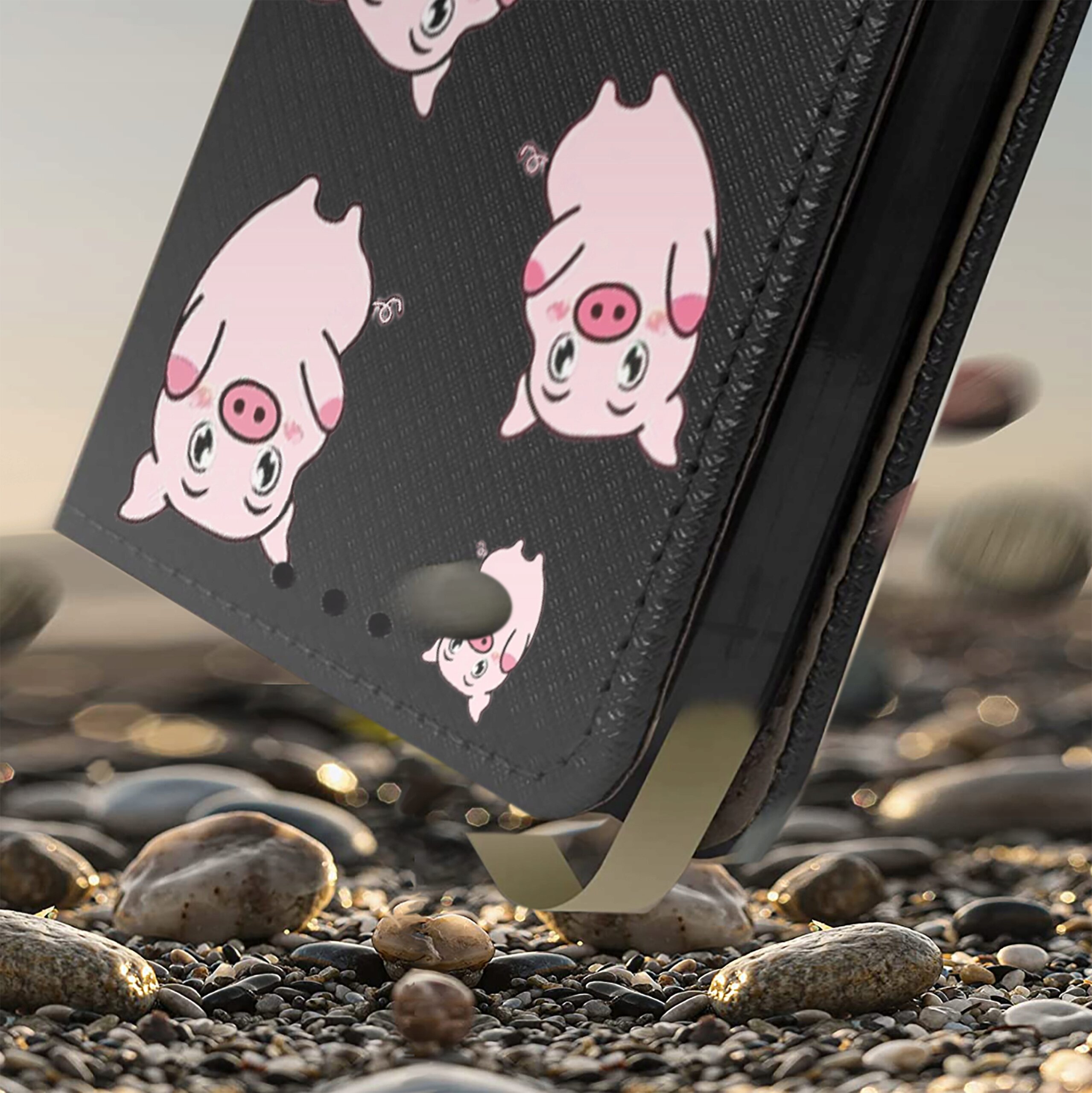 Etui na telefon z czarnym tłem ozdobione wzorem różowych świnek. Etui jest ustawione na tle kamieni.
