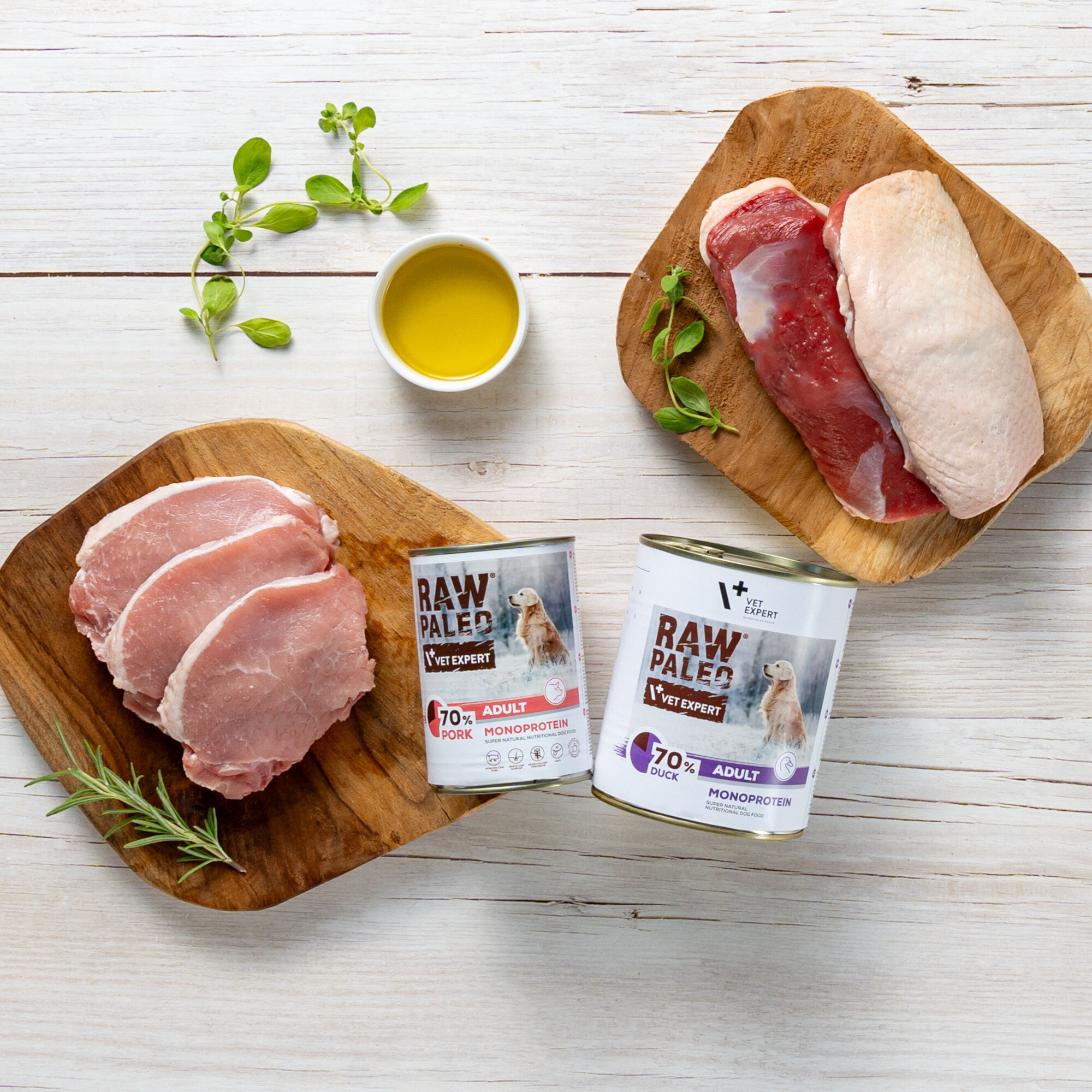 Na drewnianych deskach znajdują się kawałki surowego mięsa oraz dwie puszki karmy dla zwierząt z napisem \'RAW PALEO VET EXPERT\'. Obok puszek widoczna jest mała miseczka z oliwą i gałązki ziół.