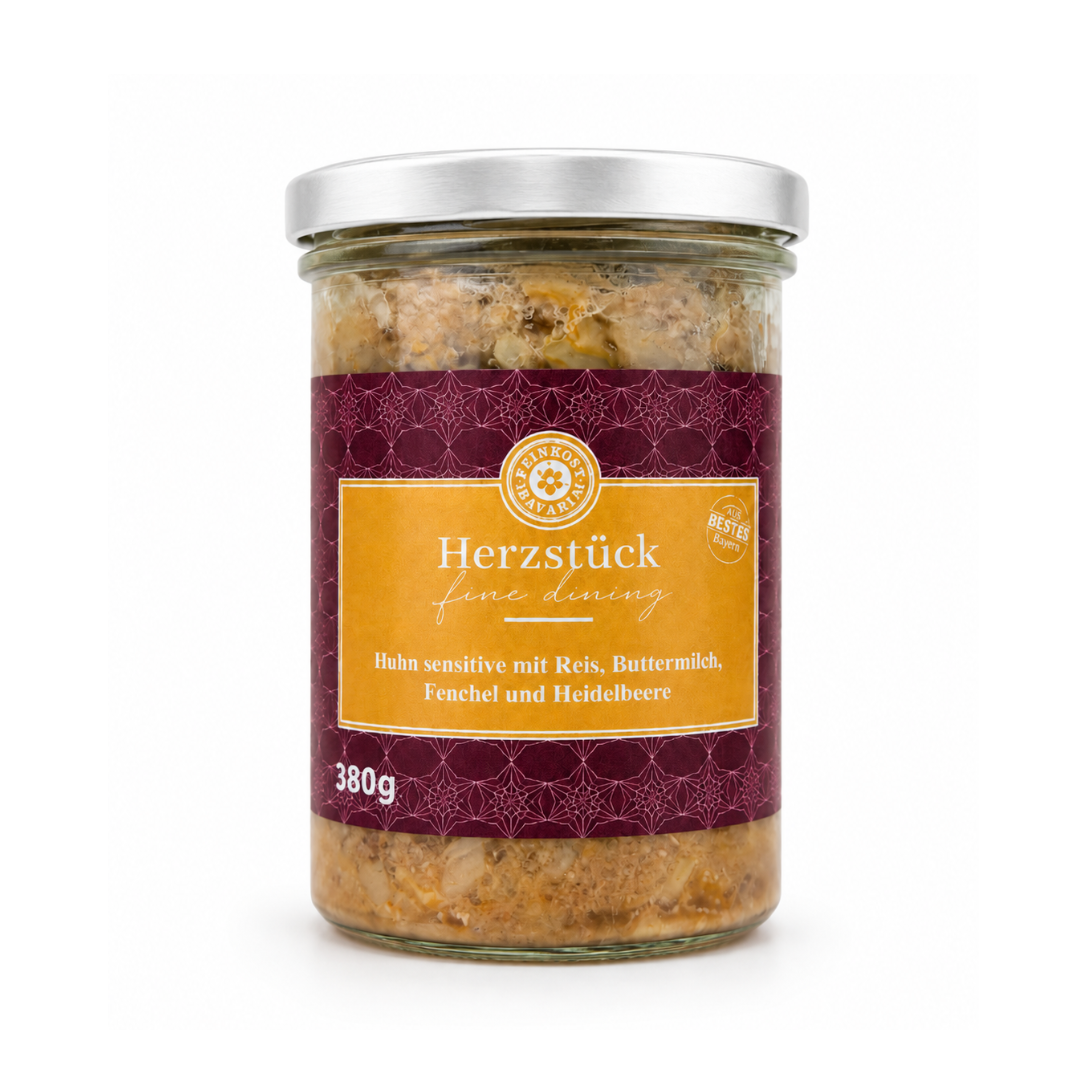 Słoik z metalową pokrywką zawiera produkt spożywczy z etykietą w kolorze bordowym i żółtym. Na etykiecie widnieje tekst: \'Herzstück fine dining Huhn sensitive mit Reis, Buttermilch, Fenchel und Heidelbeere 380g\'.
