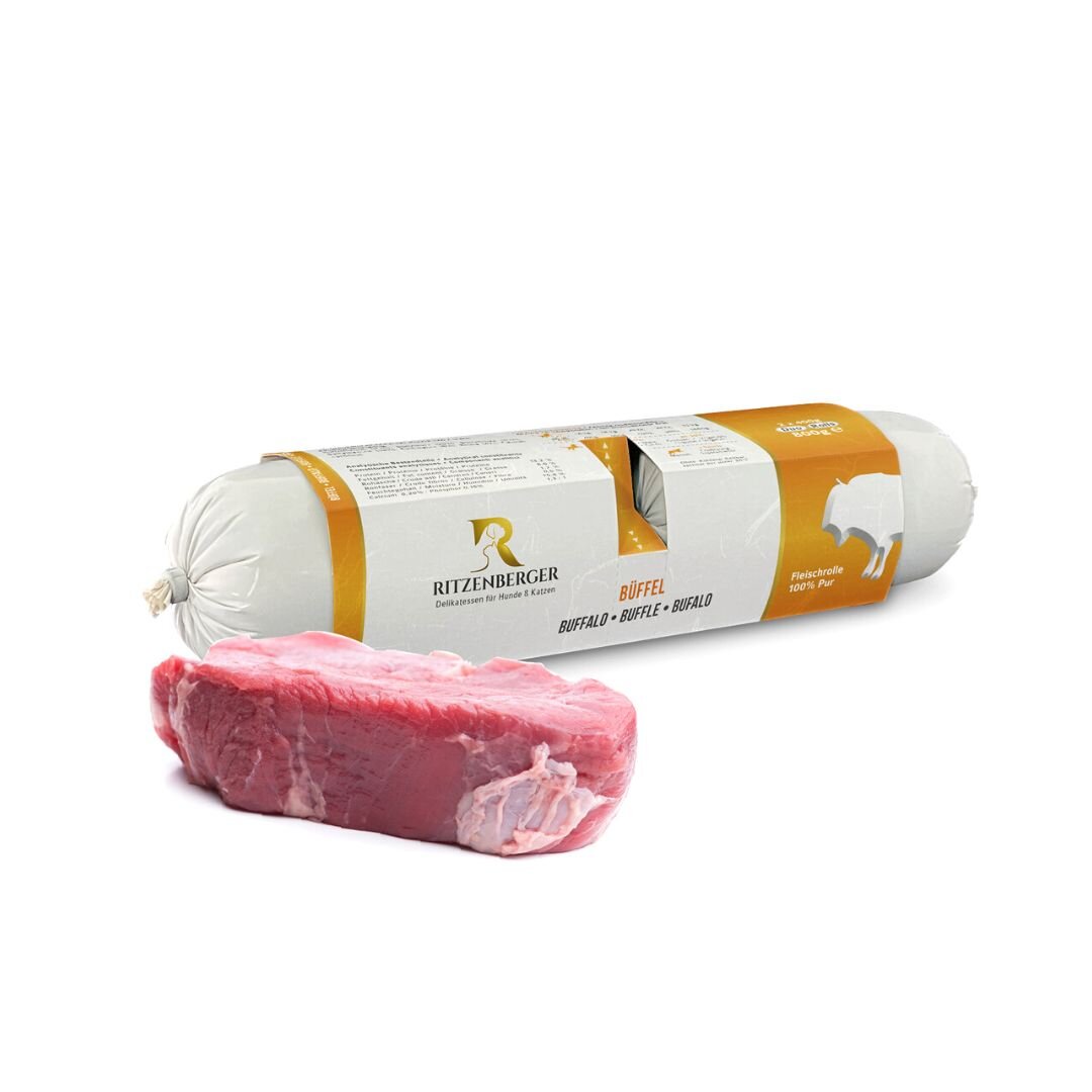 Na obrazie widoczna jest rolka karmy dla psa marki Ritzenberger z napisem \'Büffel\' oraz kawałek surowego mięsa. Opakowanie jest białe z pomarańczowym akcentem i logo producenta.