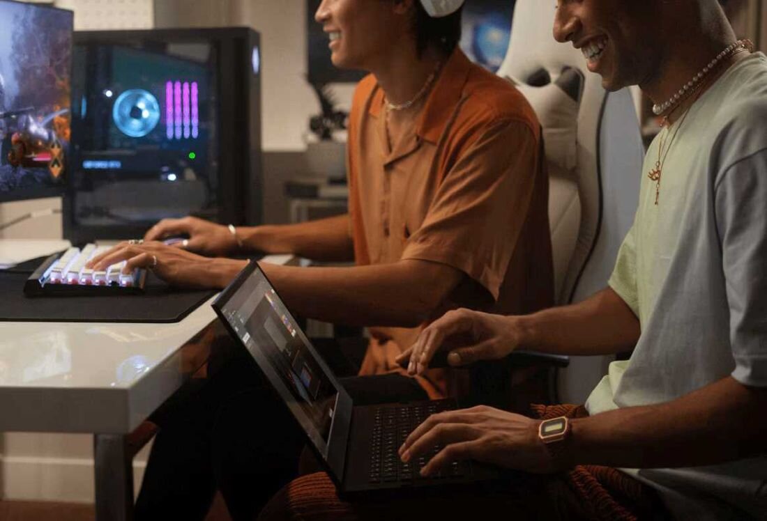 Dwóch uśmiechniętych mężczyzn siedzi przy biurku, jeden z nich używa laptopa, a drugi gra na komputerze stacjonarnym z podświetlaną klawiaturą. W tle widać monitor z grą i komputer z podświetleniem RGB.
