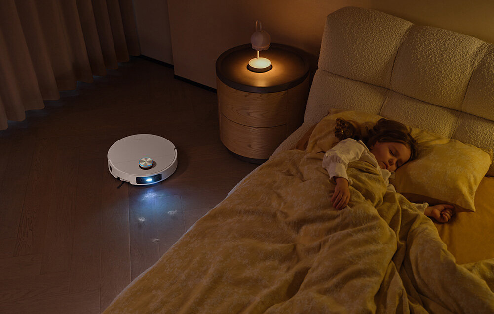 Na podłodze w sypialni znajduje się biały robot sprzątający z włączonym światłem. Obok łóżka, na którym śpi dziecko, stoi okrągły stolik z zapaloną lampką.