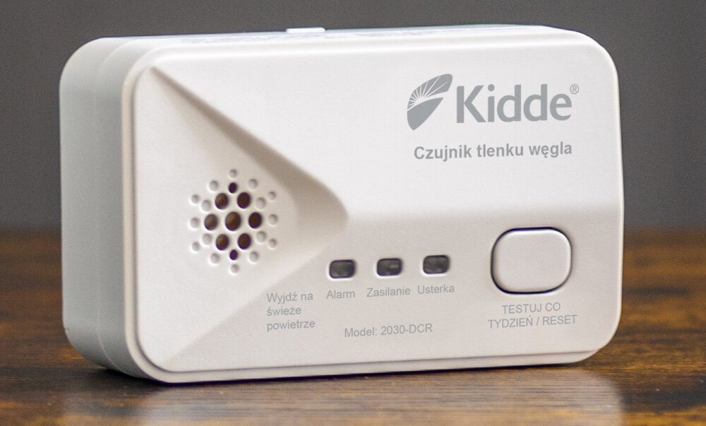 Na obrazie widoczny jest biały czujnik tlenku węgla z logo \'Kidde\' i napisem \'Czujnik tlenku węgla\'. Poniżej znajdują się przyciski i wskaźniki z opisami: \'Wyjdź na świeże powietrze\', \'Alarm\', \'Zasilanie\', \'Usterka\', \'Model: 2030-DCR\', oraz przycisk \'TESTUJ CO TYDZIEŃ / RESET\'.
