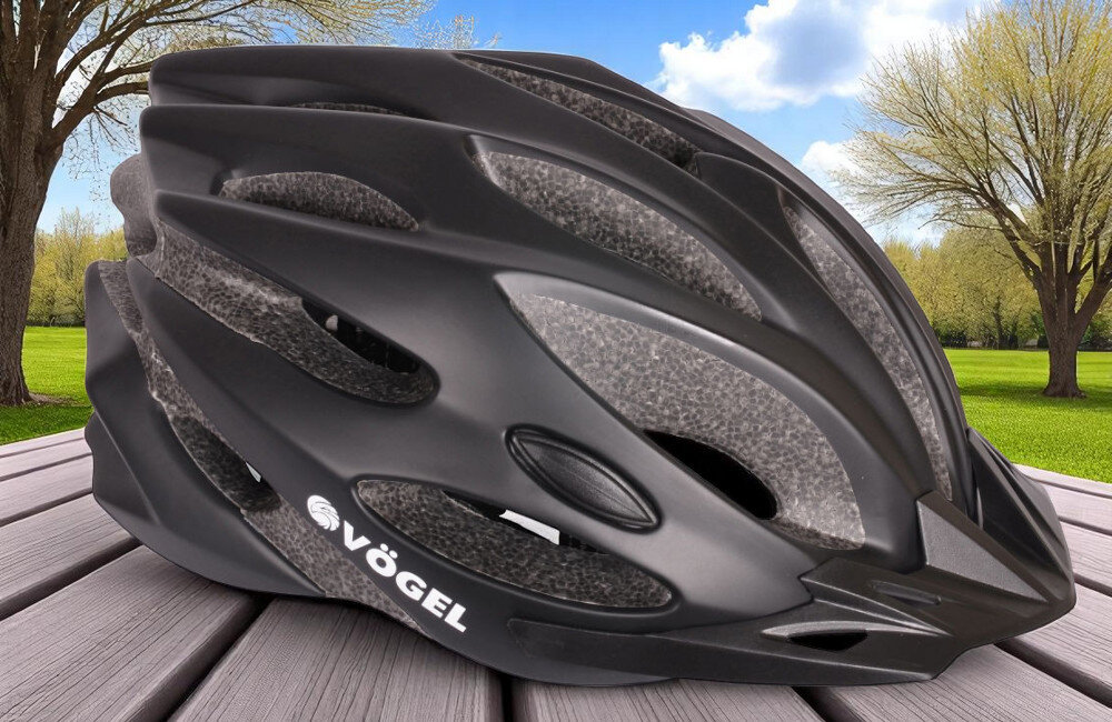 Czarny kask rowerowy z widocznym logo \'VÖGEL\' leży na drewnianej powierzchni, w tle widać zielony trawnik i drzewa pod błękitnym niebem.