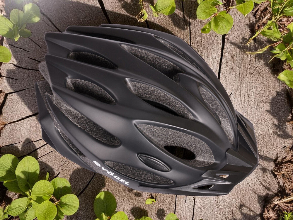 Czarny kask rowerowy z licznymi otworami wentylacyjnymi leży na drewnianej powierzchni otoczonej zielonymi liśćmi. Na kasku widoczne jest logo \'VÖGEL\'.