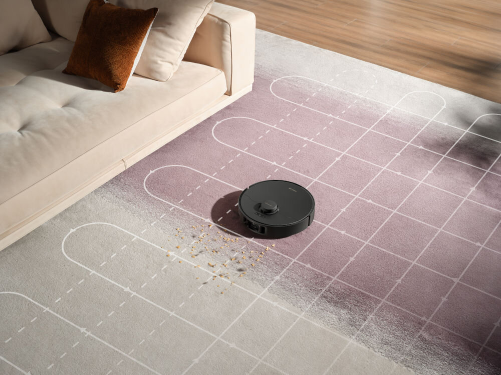 Na dywanie z geometrycznym wzorem znajduje się czarny robot sprzątający, obok którego leżą rozsypane okruchy. W tle widoczna jest jasna sofa z brązową poduszką.