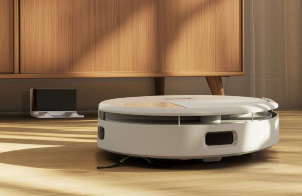 Na drewnianej podłodze znajduje się biały robot sprzątający o okrągłym kształcie. W tle widoczna jest drewniana szafka oraz stacja dokująca.