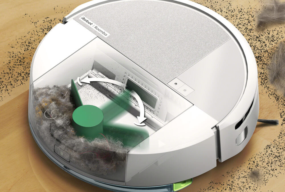 Biały robot sprzątający z widocznym logo \'iRobot Roomba\' na górnej części obudowy, otoczony przezroczystą osłoną ukazującą wnętrze z mechanizmem czyszczącym. Na podłodze wokół robota widoczne są drobinki kurzu i sierści.