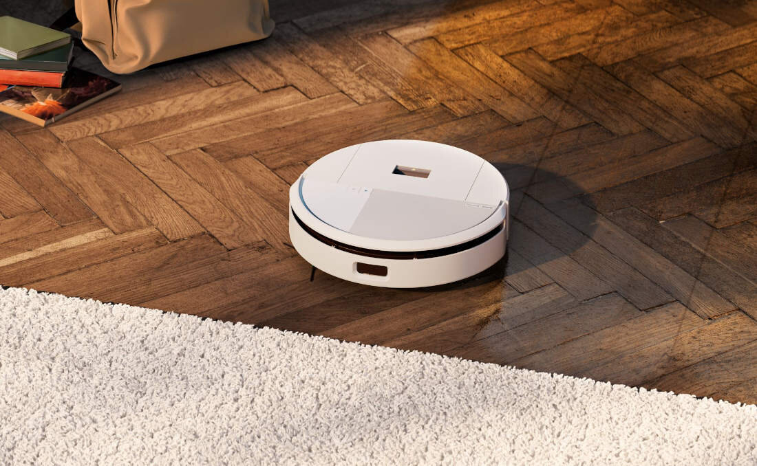 Na drewnianej podłodze znajduje się biały robot sprzątający o okrągłym kształcie, obok dywanu.