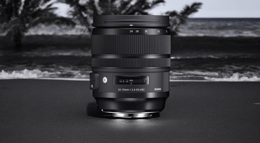 Obiektyw fotograficzny z widocznymi oznaczeniami 24-70mm 1:2.8 DG OS HSM i logo SIGMA, ustawiony na ciemnej powierzchni z rozmytym tłem przedstawiającym plażę i morze.