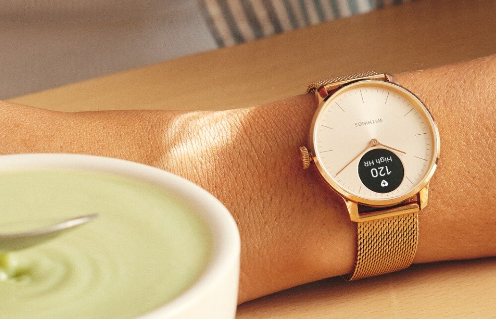 Osoba nosi różowo-złoty smartwatch z okrągłą tarczą, na której widnieje napis \'WITHINGS\' oraz wyświetlacz pokazujący \'120 HIGH HR\'. Obok znajduje się miska z zielonym płynem i łyżką.