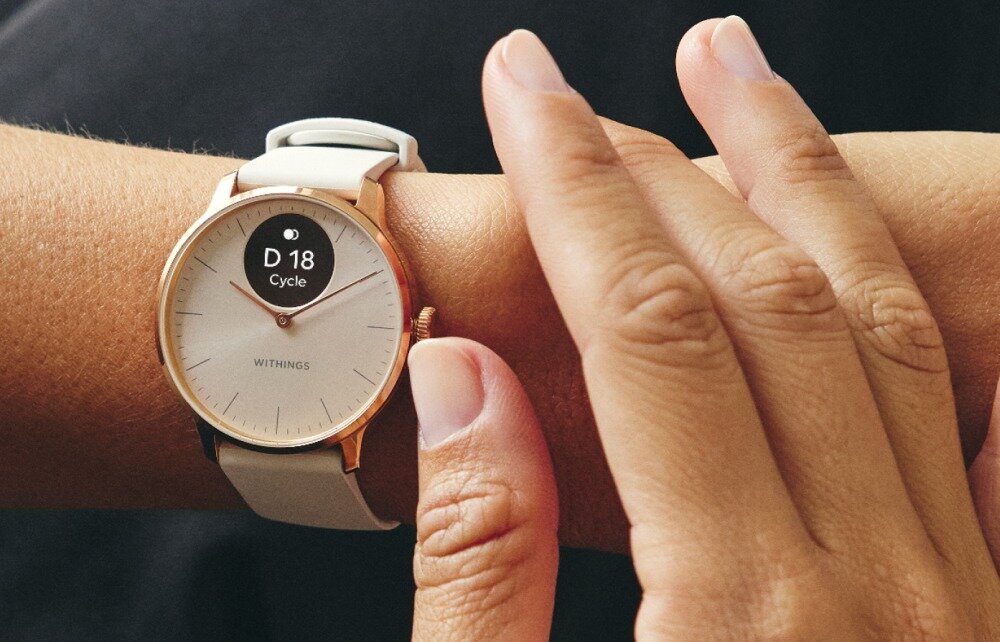 Na zdjęciu widoczny jest smartwatch z okrągłą tarczą w kolorze różowo-złotym, z wyświetlaczem pokazującym \'D 18 Cycle\'. Na tarczy znajduje się napis \'WITHINGS\'.