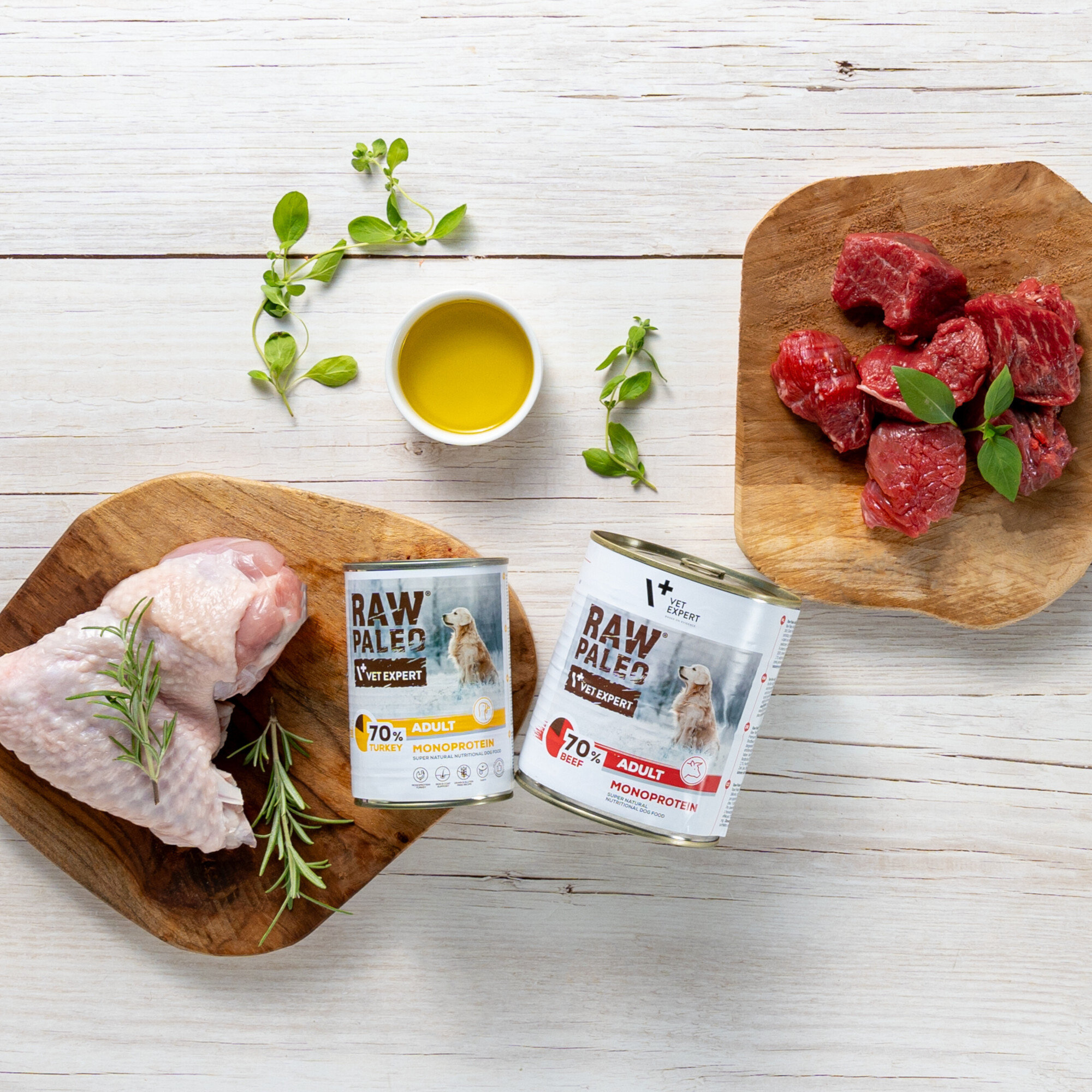 Na drewnianym tle znajdują się dwa opakowania karmy RAW PALEO VET EXPERT, jedno z indykiem, drugie z wołowiną, obok surowe mięso i zioła. Widoczna jest także miseczka z oliwą.